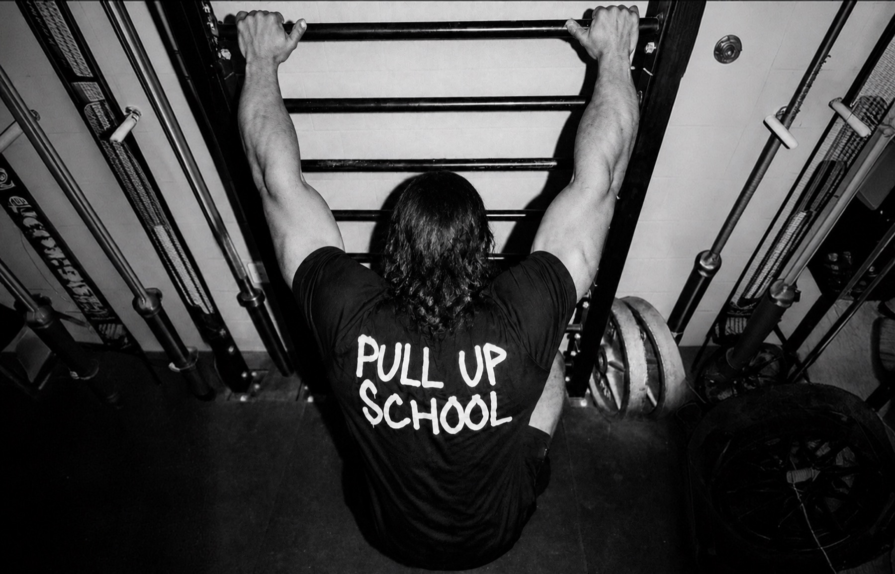 Pulling up to the bar in a Pull Up School shirt, seen from behind